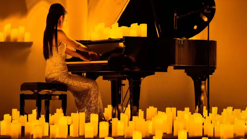 A pianist in a shimmering gold dress performs on a grand piano, surrounded by glowing candlelight. The warm, golden ambiance enhances the intimate and elegant atmosphere of the scene.